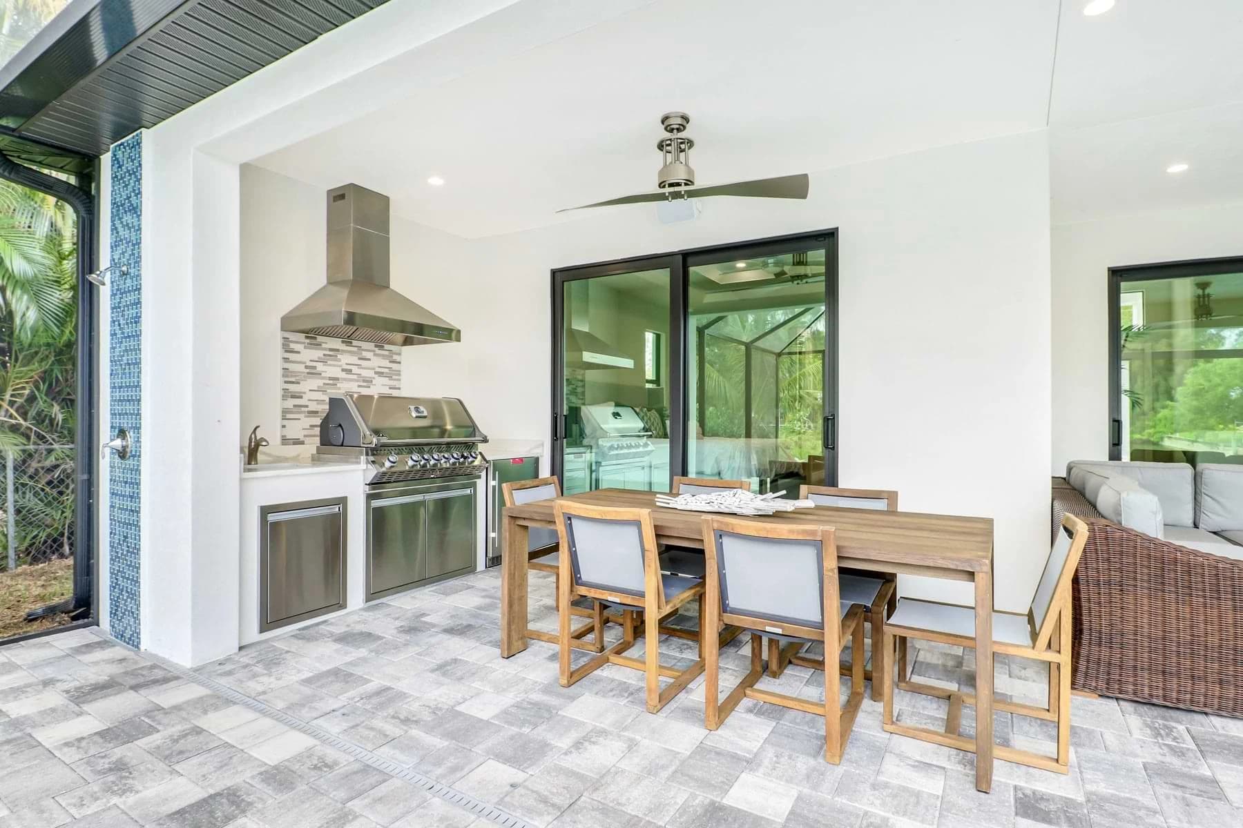 Spica poolside covered lanai with a stainless steel outdoor kitchen, tiled backsplash, wood dining table, and wicker lounge seating