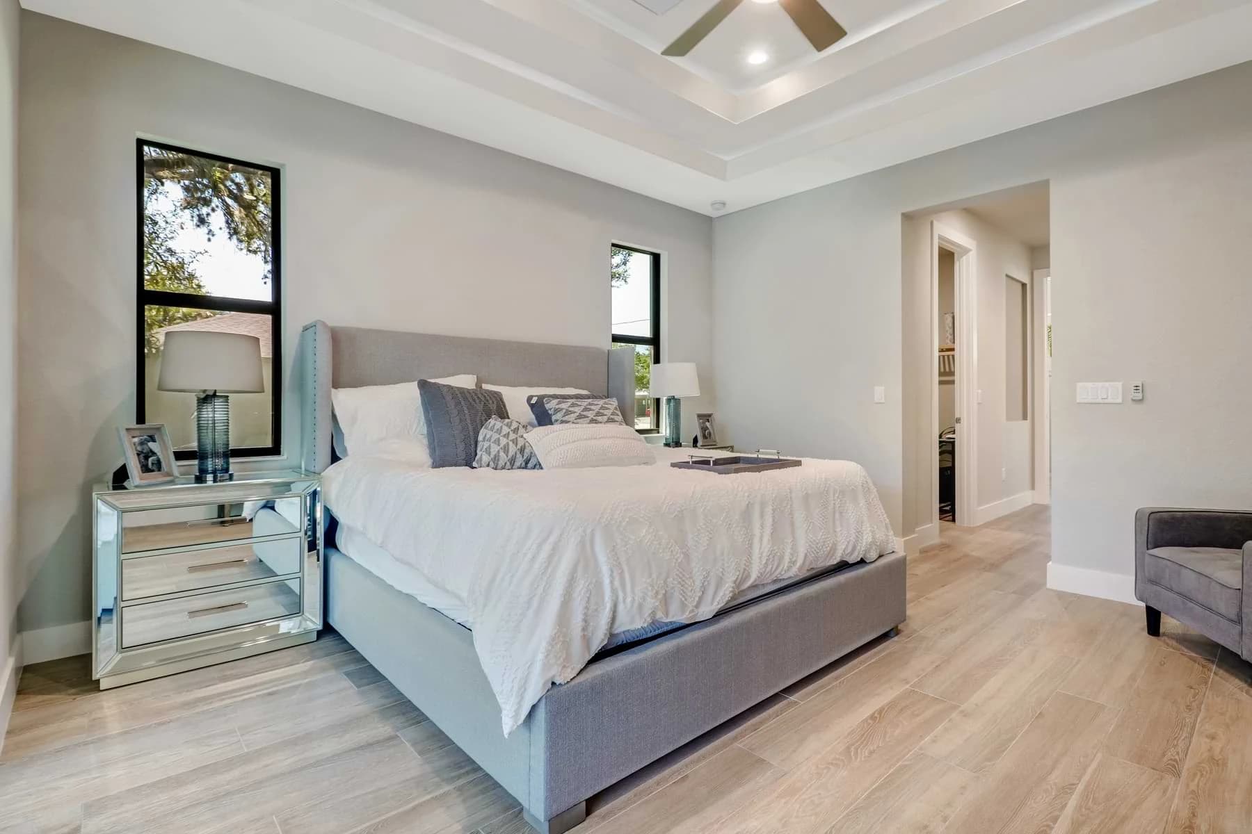 Spica furnished bedroom with a gray upholstered bed, mirrored nightstands, tall black-framed windows, and layered tray ceiling