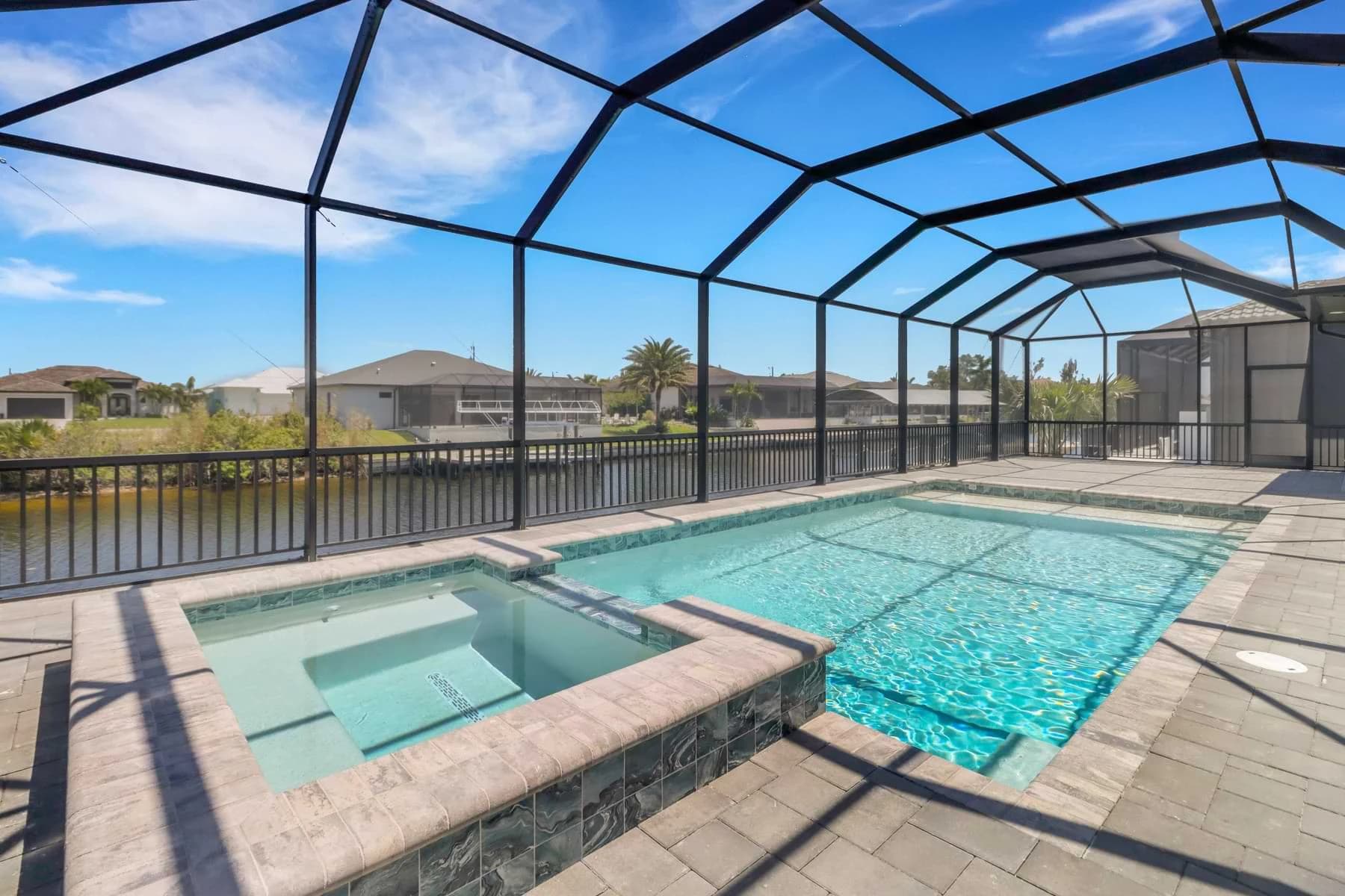 Vista Grande screened canalfront pool with a raised stone-look tiled spa, spillover edge, and beige paver surround