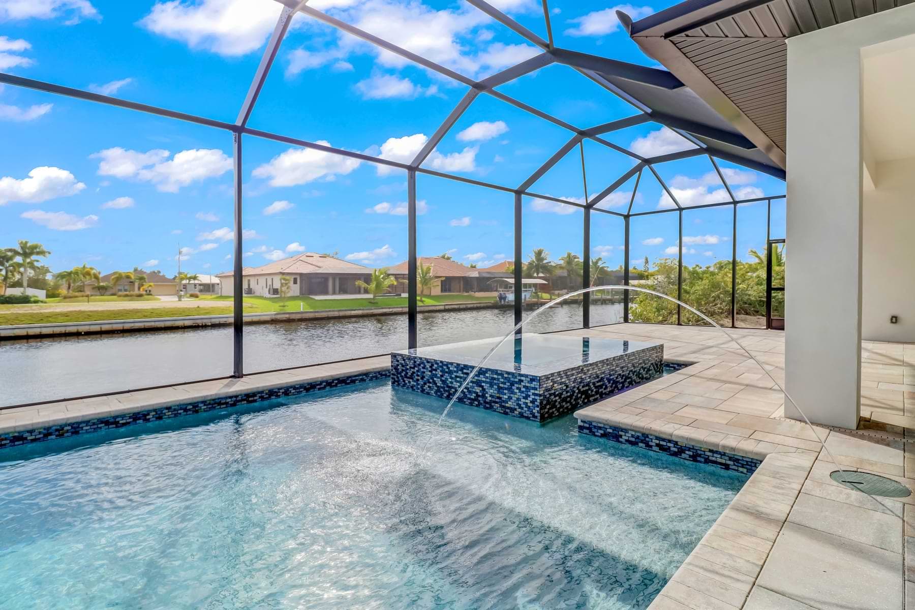 Mira Vista screened canalfront pool with a raised blue mosaic spa, arcing water jets, and pale paver deck