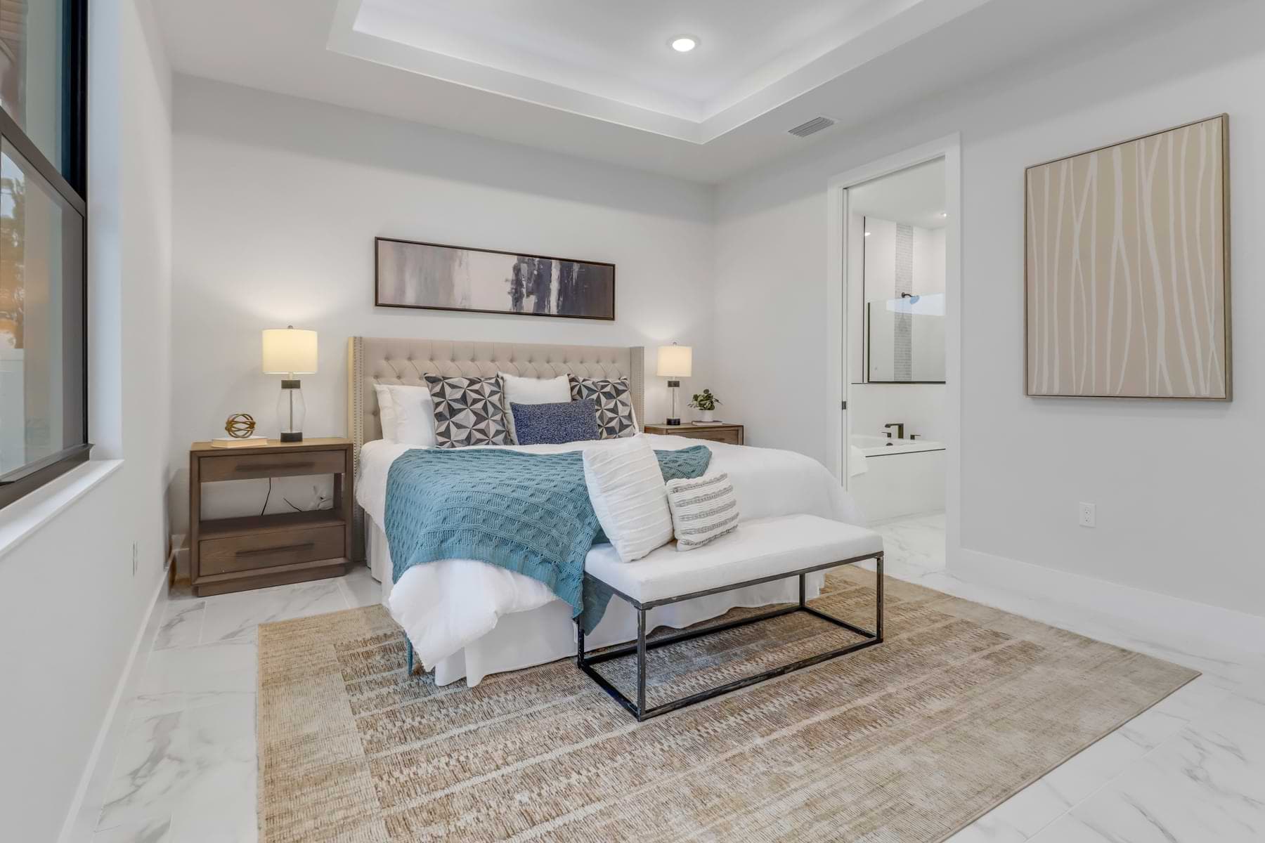 Hardwyn furnished bedroom with an upholstered headboard, blue bedding, bedside lamps, bench, and illuminated tray ceiling