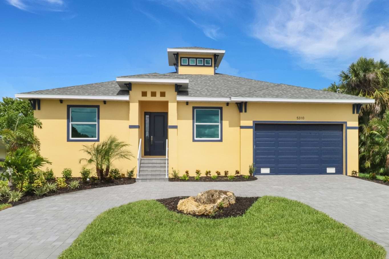 Front exterior of a yellow Key West home with a dark garage door, raised entry, and paver driveway