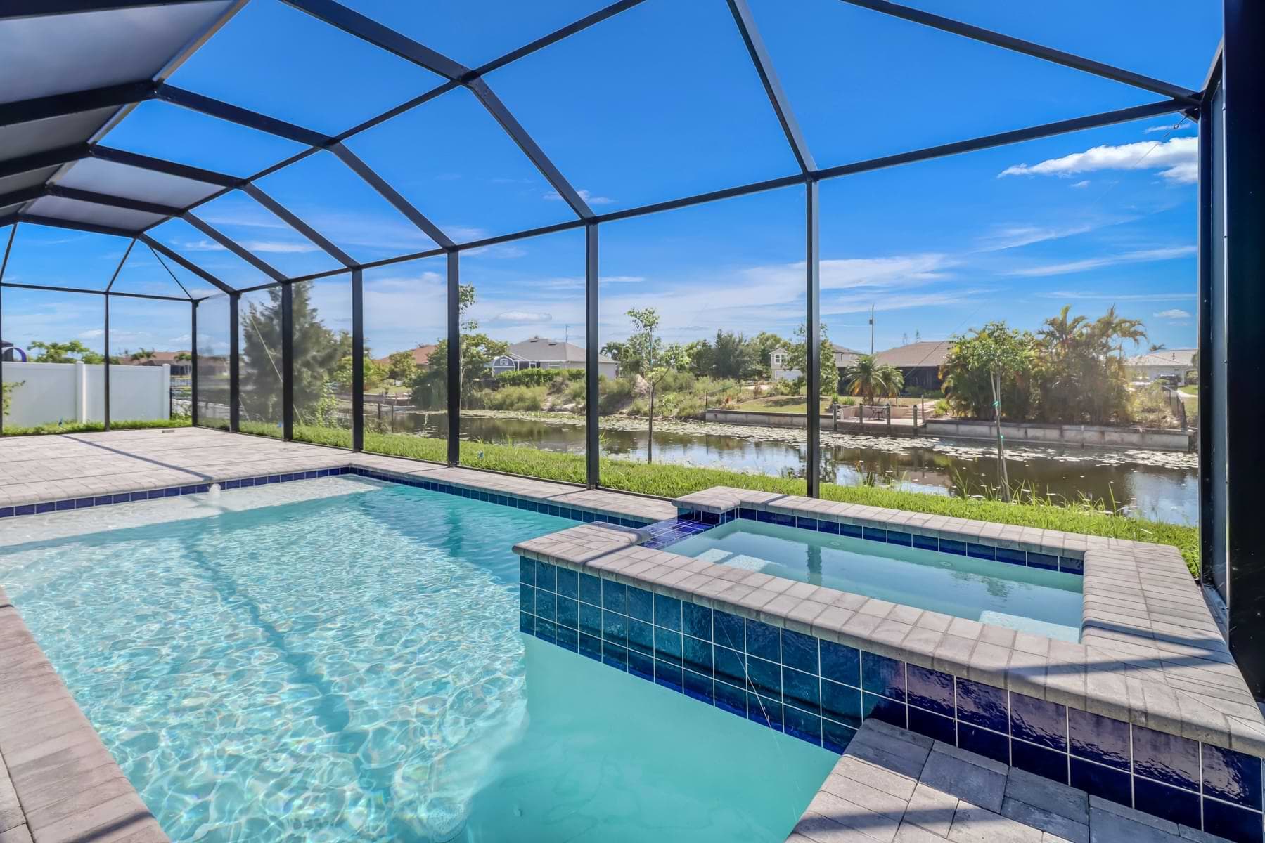 Catalina screened canalfront pool with a raised blue-tiled spillover spa and pale paver deck in bright sunlight