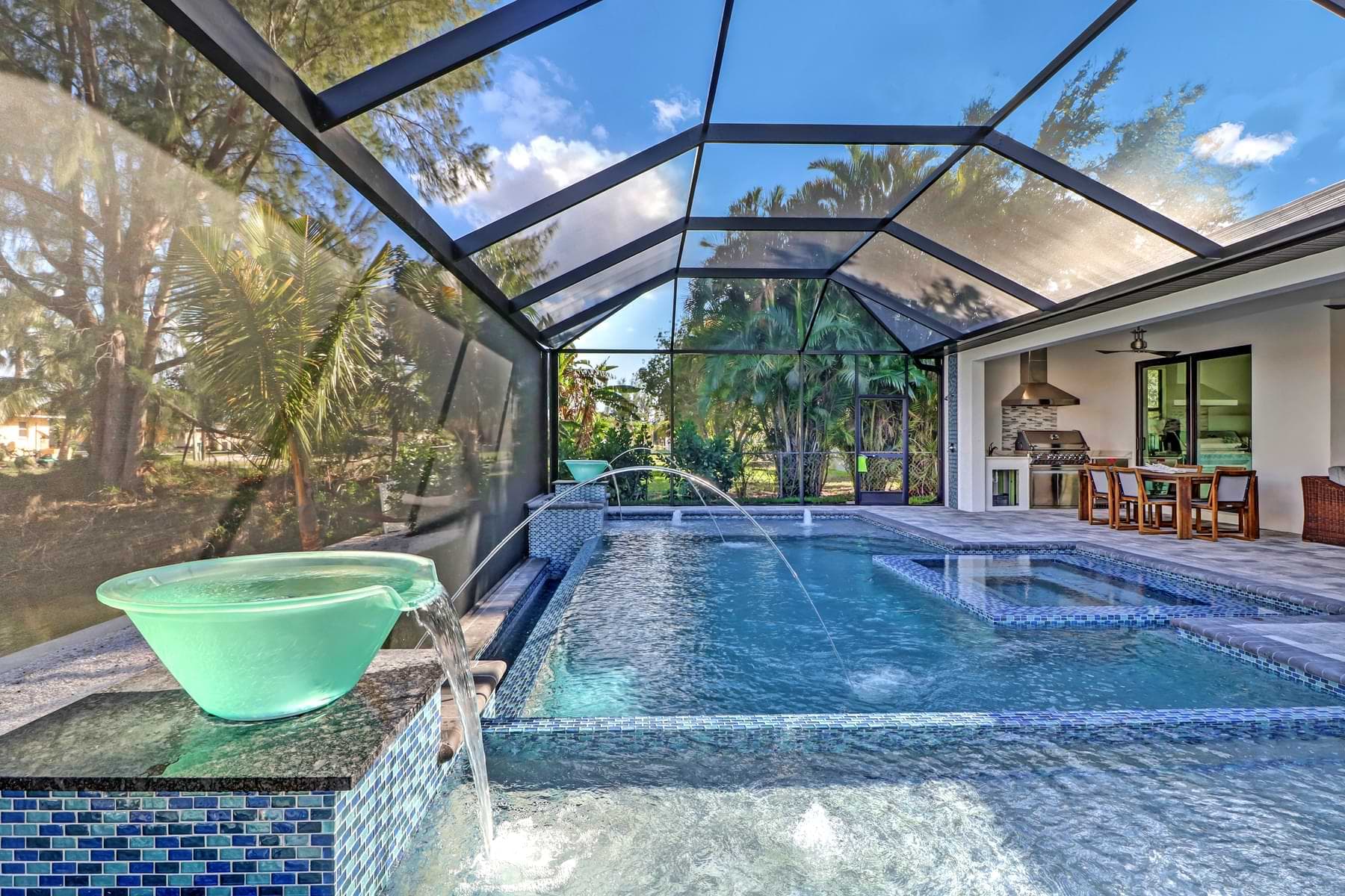 Spica screened pool with glass bowl waterfalls, arcing water jets, blue mosaic tile, and a furnished outdoor kitchen