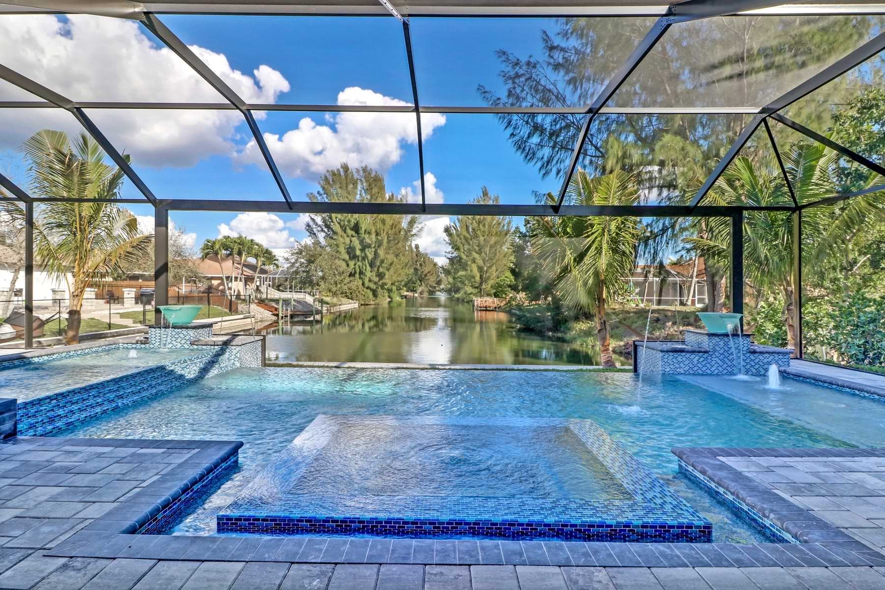 Screened Spica pool with a blue-tiled sun shelf and glass bowl waterfalls overlooking a canal