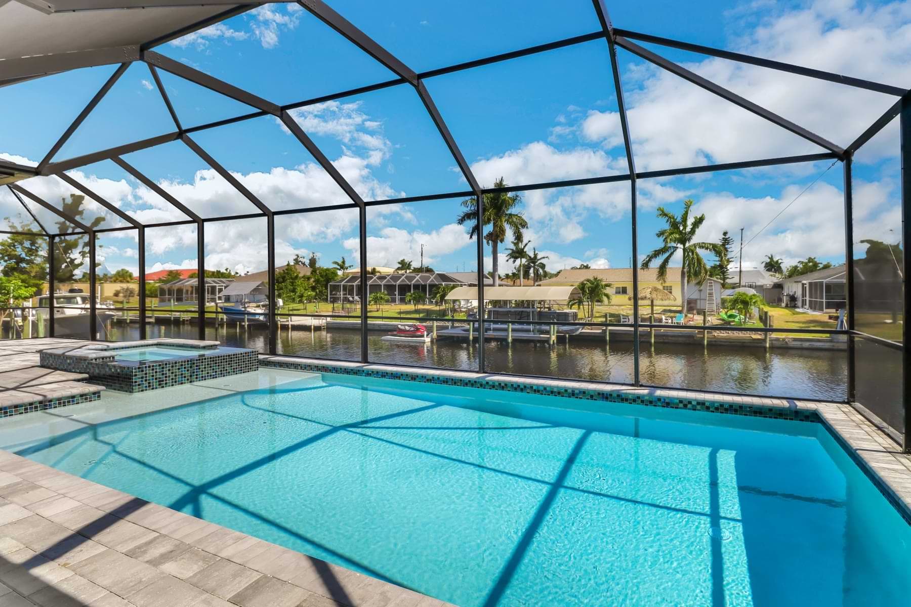 Mira Vista screened pool with a broad shallow sun shelf, raised mosaic spa, and canalfront palms and boat docks