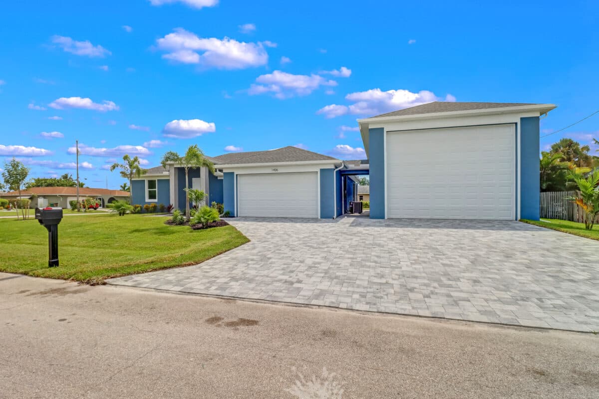 Front exterior of a blue home with white garage doors, a landscaped lawn, and a wide paver driveway